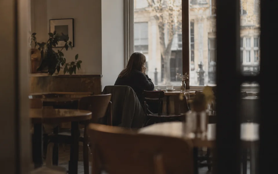 A woman sitting alone at a cafe table by a window, seen from across the room through an open doorway, half turned away.